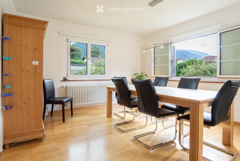 Bright dining area with wooden floor, large windows offering a scenic view, a dining table surrounded by black chairs, and a wooden wardrobe.