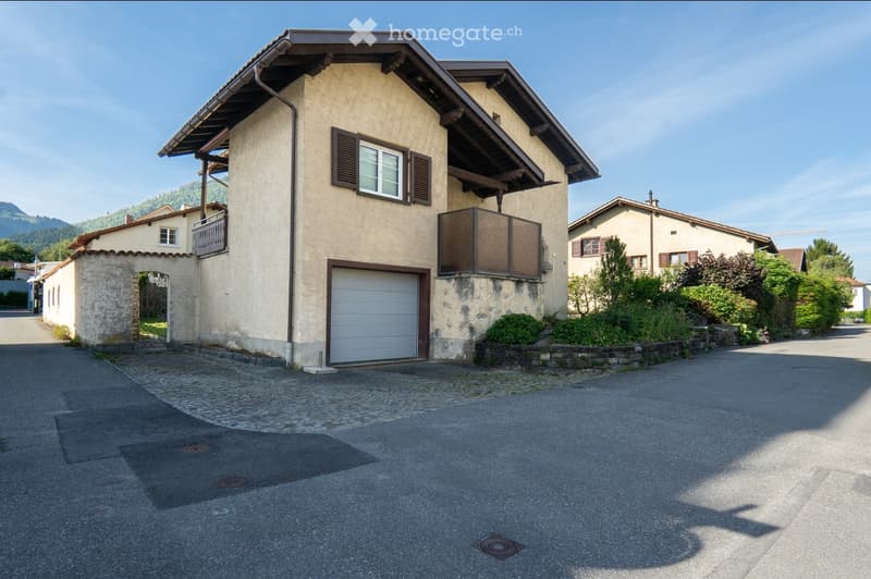 Two-story house with beige facade, wooden shutters, garage, and balcony. Surrounded by greenery and a clear view of mountains in the background.