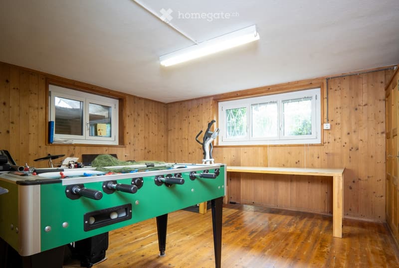 A room with wooden walls and flooring, featuring a foosball table, a long wooden table, an exercise bike, and a drum set in the background.