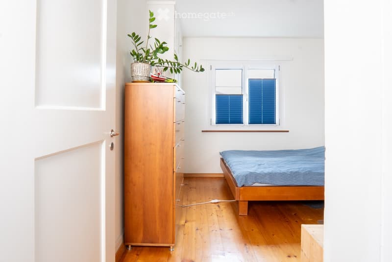 Minimalist bedroom with wooden floor, single bed with blue bedding, tall wooden wardrobe, small plant on top, and a window with blue shutters.