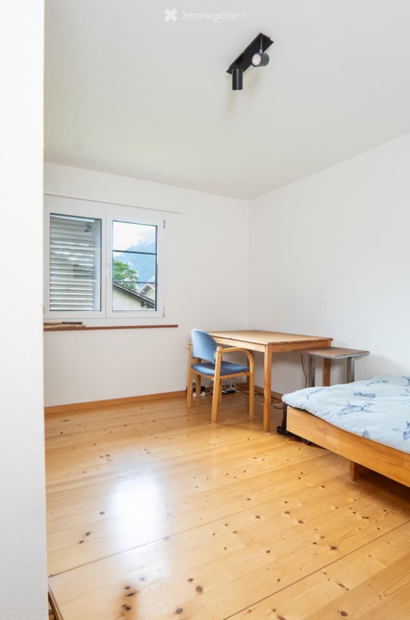 Bright, minimalist bedroom with wooden flooring, white walls, and large windows offering a scenic view. Features a single bed, a wooden desk with a chair, and a small stool.