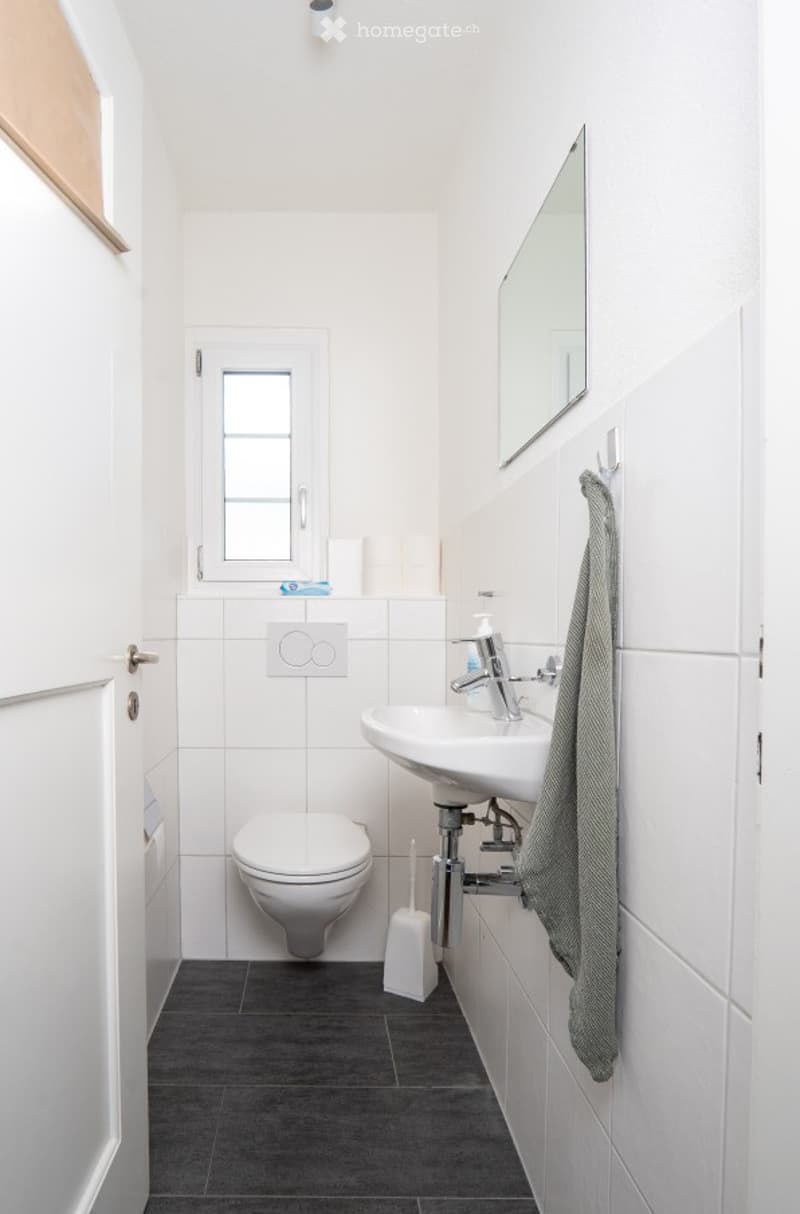 Modern, compact bathroom with white tiles, a toilet, a sink with a mirror, a towel, and a small window for natural light.