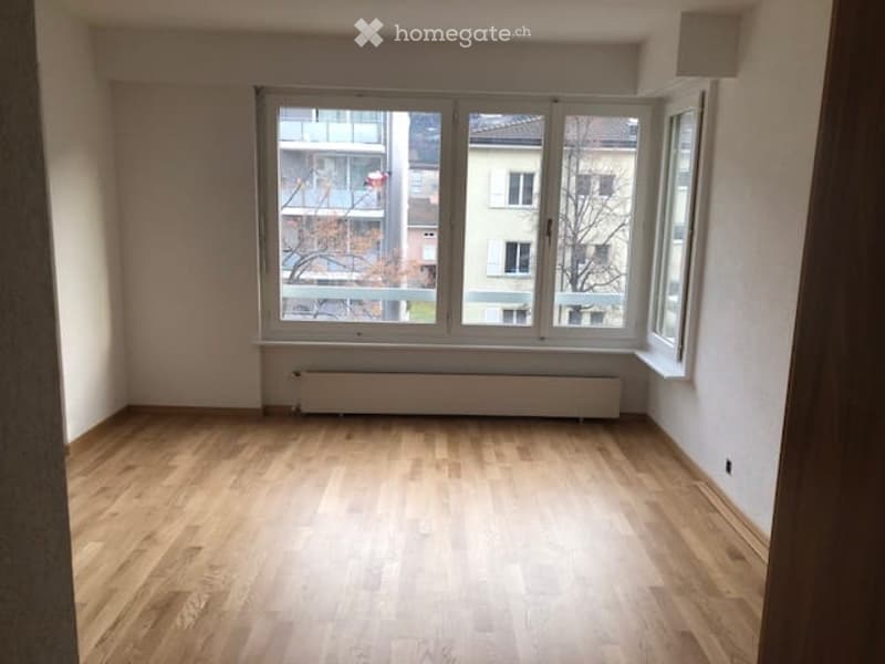 Empty room with wooden flooring, large window, radiator, and white walls.