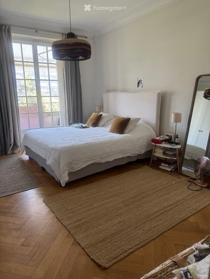 Cozy bedroom with a large bed, nightstand, mirror, and a fan. The room features wooden flooring, a large window with curtains, and a woven rug.