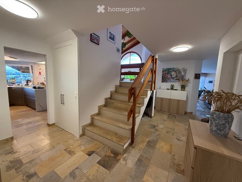 Spacious hallway with tiled flooring, wooden staircase with a handrail, and a view into the kitchen and living area. Features modern lighting and decorative elements.