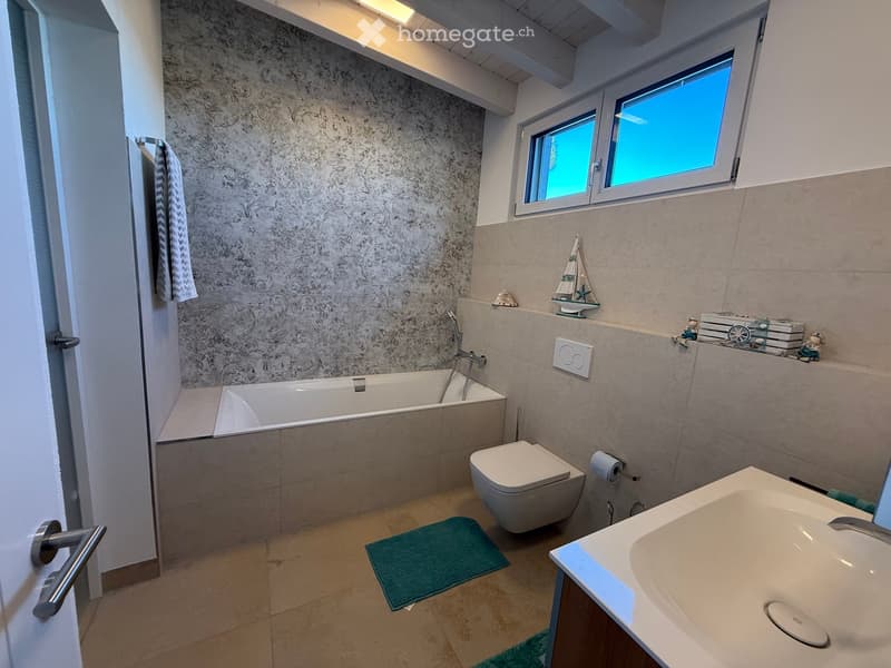 Modern bathroom with a bathtub, toilet, sink, and decorative items on a shelf, including a model boat and books.