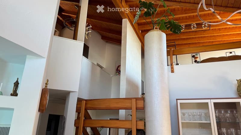 Stylish living room with wooden ceiling beams, white walls, a tall white column, a wooden chair, a glass cabinet with wine glasses, and a decorative vase with a leafy plant.