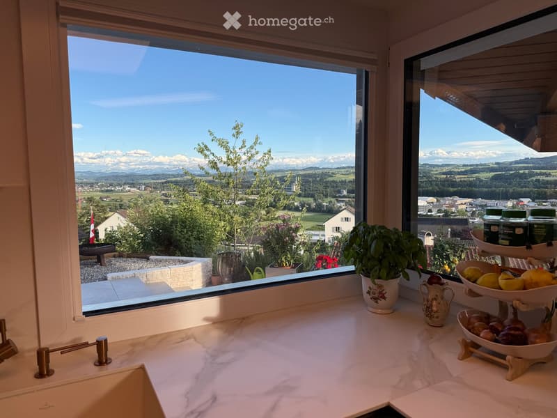 Kitchen with marble countertops, sink, and decorative bowls filled with fruits and vegetables, overlooking a scenic valley and mountains.