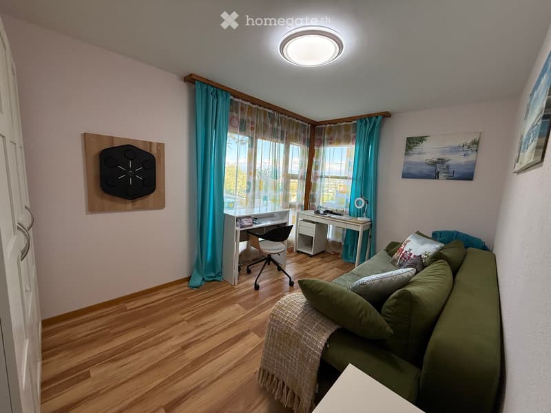 A cozy, well-lit room with a green sofa, a desk with a chair, and a small table. The room features wooden flooring, large windows with blue curtains, and a modern ceiling light.