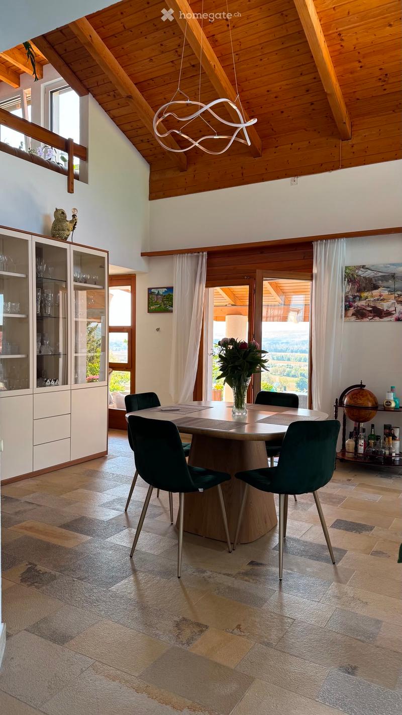 Dining area with a round wooden table, six black chairs, a modern pendant light, a glass cabinet, and a vase with flowers on the table.