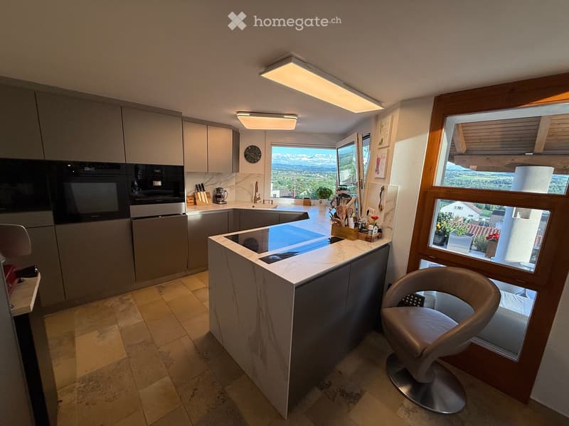 Modern kitchen with grey cabinets, marble countertop, built-in oven, dishwasher, and a view of the landscape through large windows.