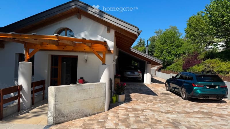 Two-story house with a wooden roof, white walls, and a covered entrance. Garage with two cars parked inside and outside, surrounded by greenery.