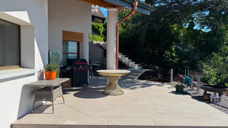 Outdoor patio area with a round table, wicker chair, barbecue, potted plants, and steps leading up to a raised garden area.