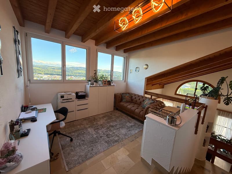 Bright, multi-functional room with exposed wooden beams, large windows offering scenic views, a comfortable sofa, a desk with office supplies, and a small kitchen area with a cabinet and teapot.
