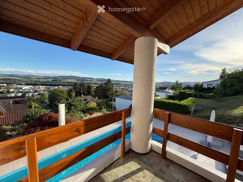 Balcony with wooden railings, tiled floor, and scenic view of a town and surrounding landscape.
