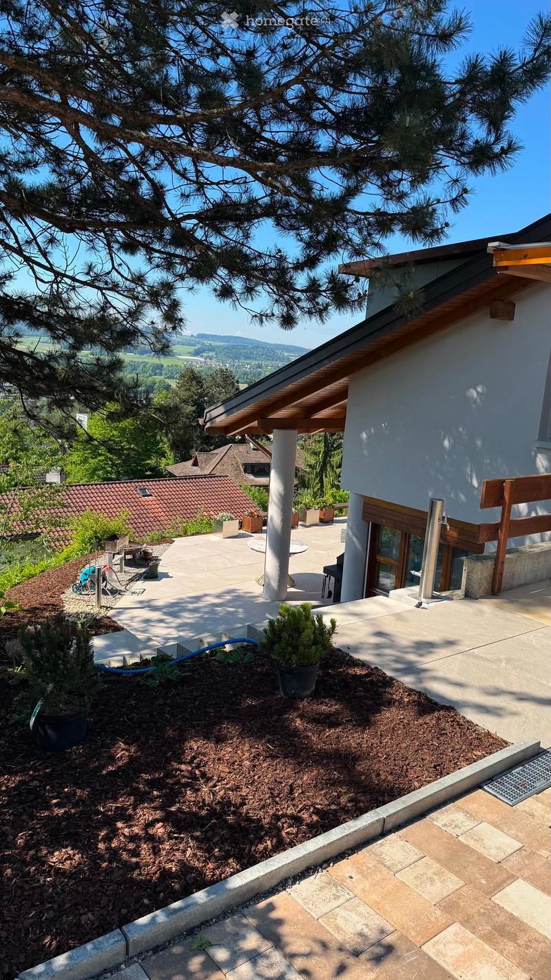 A modern outdoor patio area with a wooden pergola, potted plants, and a scenic view of a valley and mountains.