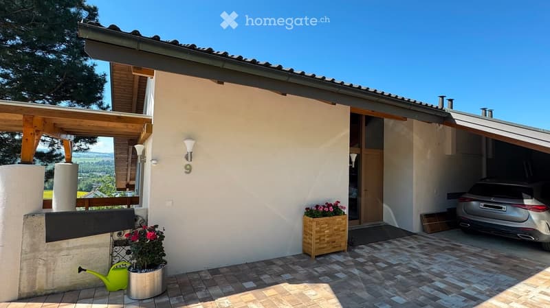 A modern two-story house with a garage, a covered patio area, and a scenic view of the countryside. The house features a light-colored exterior, wooden beams, and decorative flower boxes.