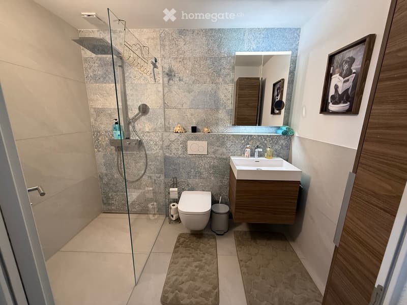 Modern bathroom with a glass-enclosed shower, a white toilet, a wooden vanity with a sink, and a mirror above the sink. The walls are tiled in a grey marble pattern, and the floor is tiled in beige.