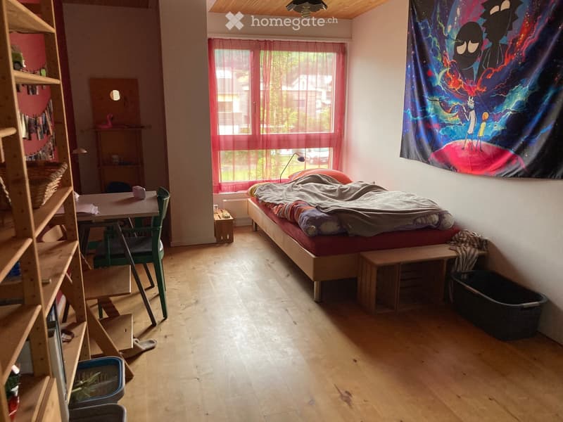 Cozy bedroom with wooden floor, bed with red mattress, small table with chair, shelf with books and basket, and a colorful tapestry on the wall.