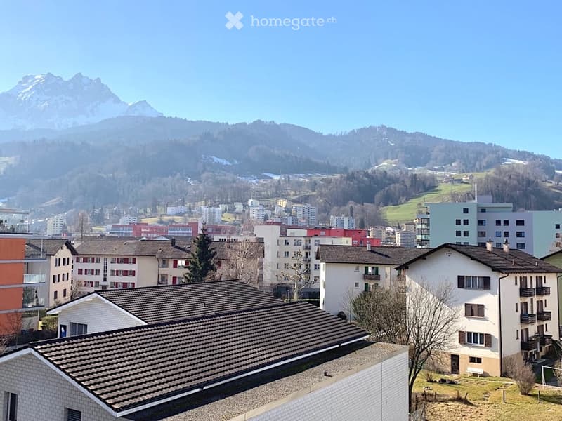 Panoramic view of a town with residential buildings, surrounded by green hills and snow-capped mountains under a clear blue sky.