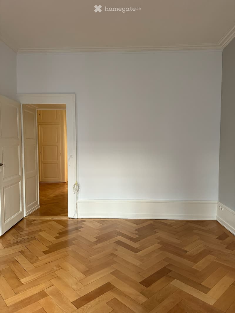Empty room with light-colored walls, wooden herringbone flooring, and an open doorway leading to another room.