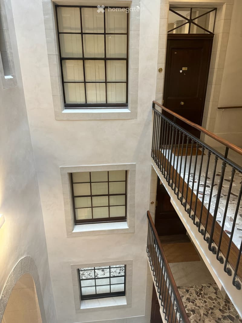 Staircase with wooden handrail and metal balusters, beige walls, and multiple windows with white frames and black grilles.