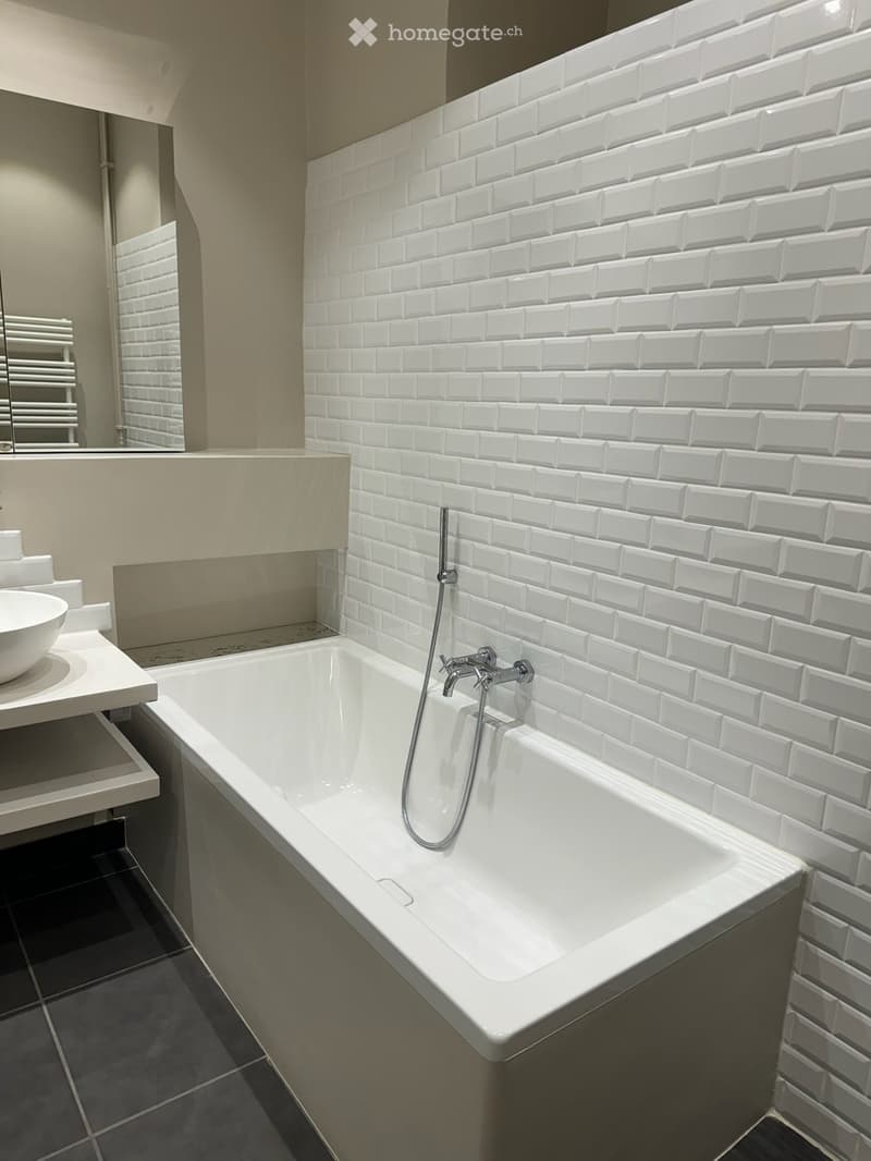 Modern bathroom with a white bathtub, chrome faucet, white sink, and a large mirror. The walls are tiled with white rectangular tiles.