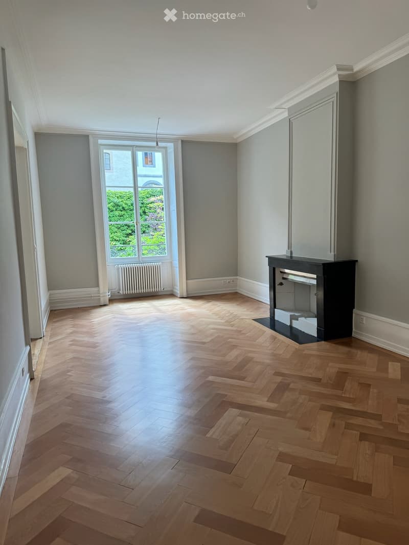 Empty room with herringbone wooden flooring, grey walls, large window with view of greenery, radiator, and fireplace.
