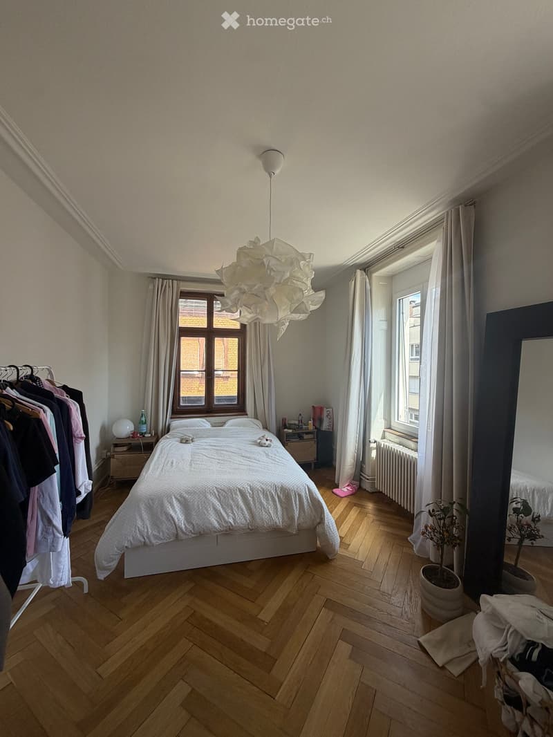 Cozy bedroom with wooden flooring, large window with curtains, bed with white bedding, nightstand with lamp, clothing rack, and potted plants.