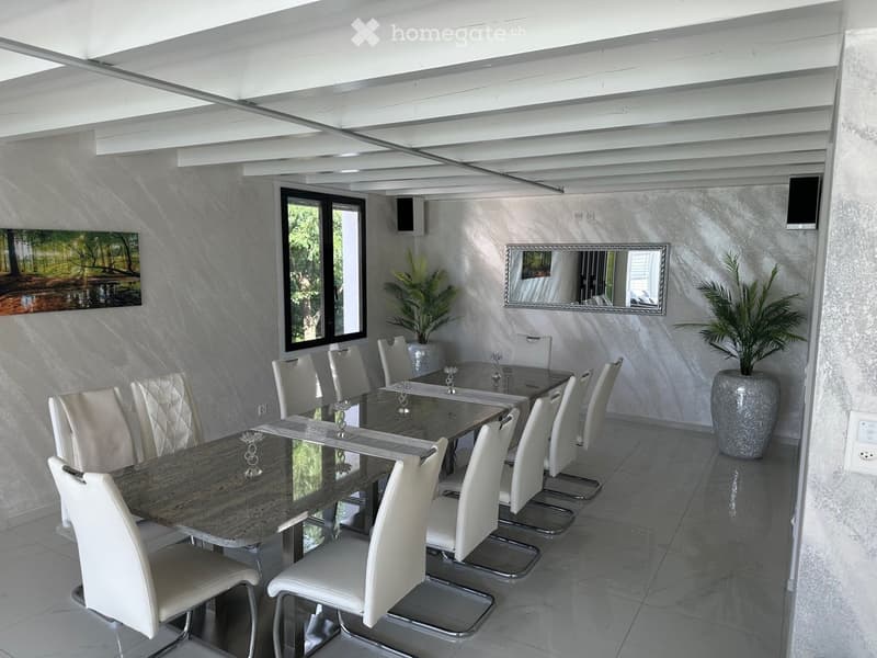 Modern dining room with a long marble dining table, white chairs, a large mirror, and decorative plants.