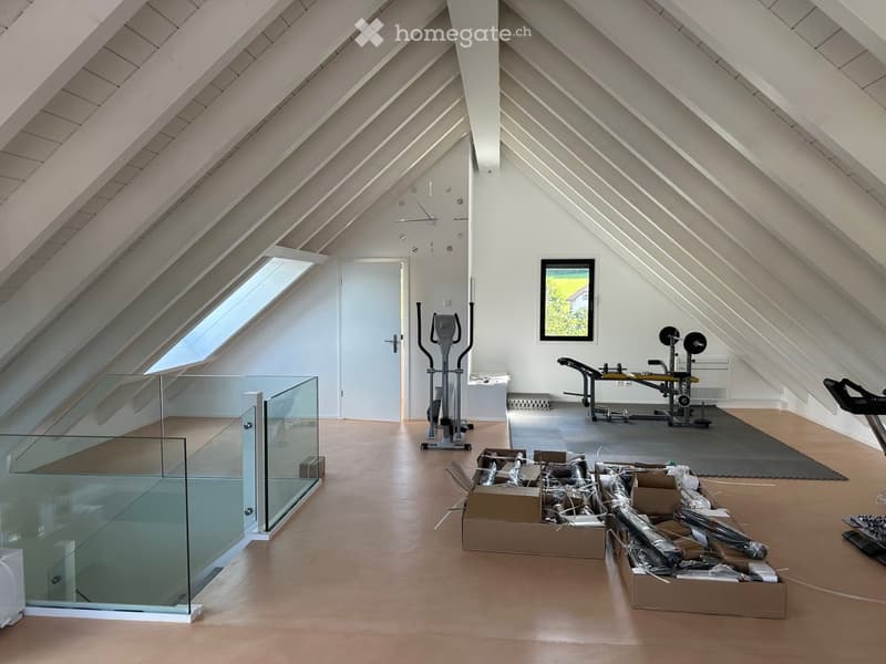 An attic space with wooden floors, slanted ceilings, and large windows. Contains exercise equipment, a glass railing, and several boxes.