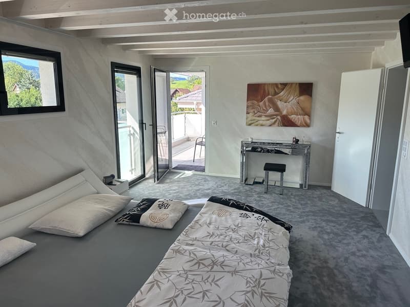 Modern bedroom with a large bed, sofa, mirror, and a view of the mountains through large windows and a balcony door.