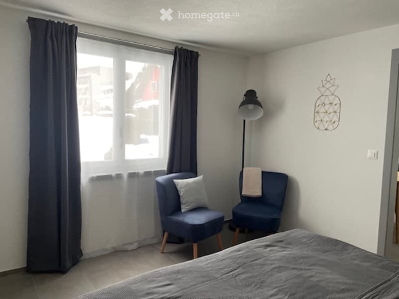 Cozy bedroom with a bed, two blue armchairs, a floor lamp, and a window with dark curtains.