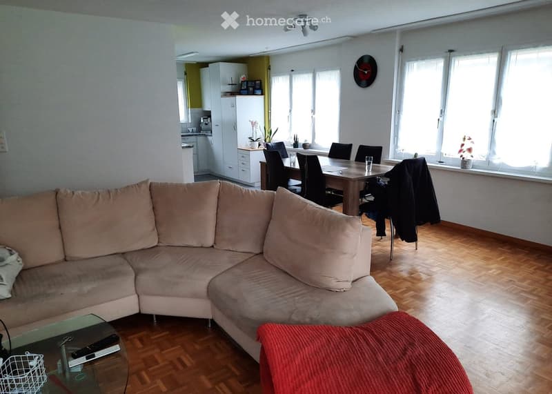 A modern living room with a large L-shaped sofa, a glass coffee table, and a dining area with a table and chairs. The room features wooden flooring, large windows, and a kitchen area visible in the background.
