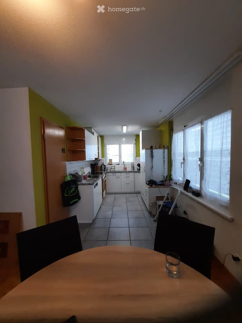 A modern kitchen with white cabinets, a tiled floor, and a central island. The space includes a dining table with a glass, a refrigerator, and various kitchen appliances.