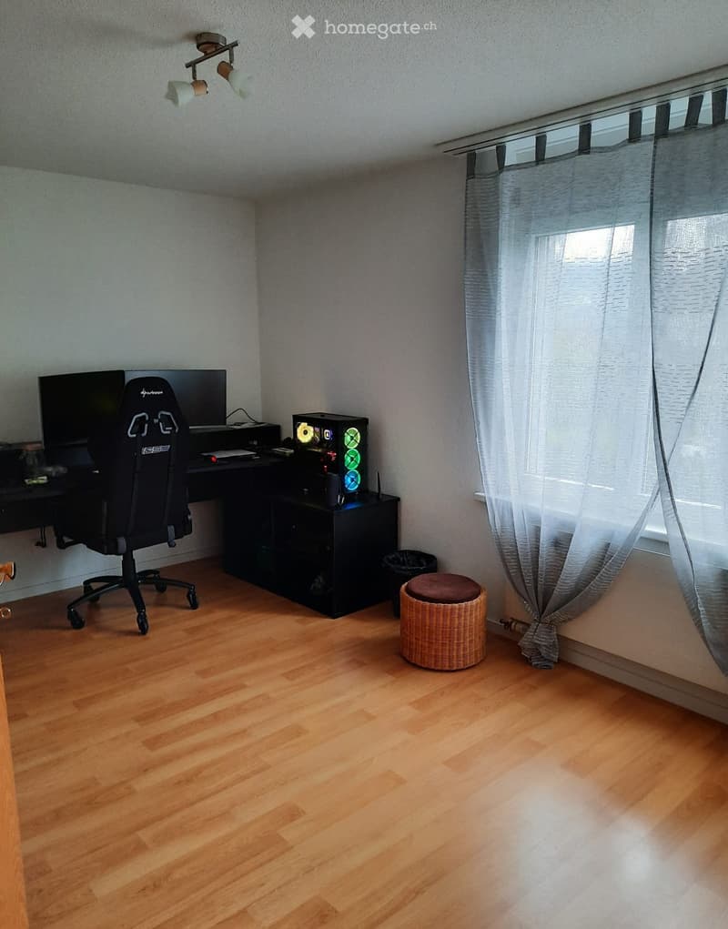A furnished room with wooden flooring, a large window with curtains, a black desk with a computer setup, a gaming chair, and a small wicker stool.