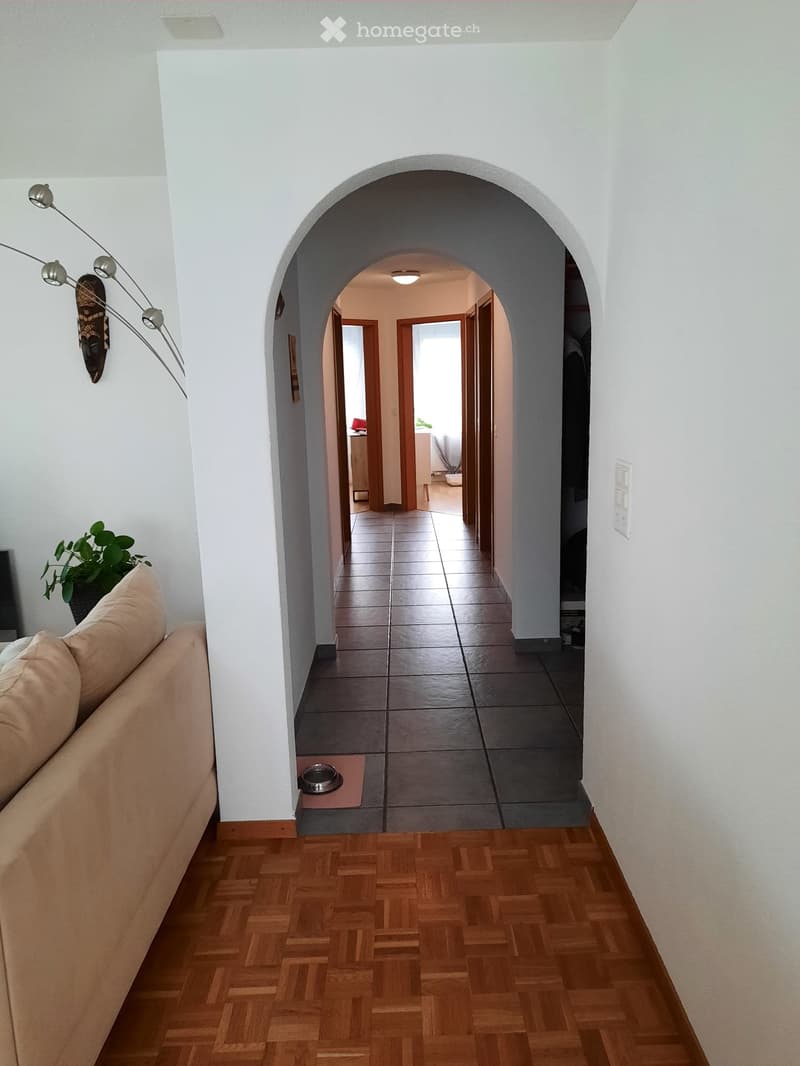 A well-lit hallway with wooden flooring, white walls, and an arched entryway leading to another room. Visible elements include a beige sofa, a decorative wall-mounted light fixture, and a small plant in a pot.