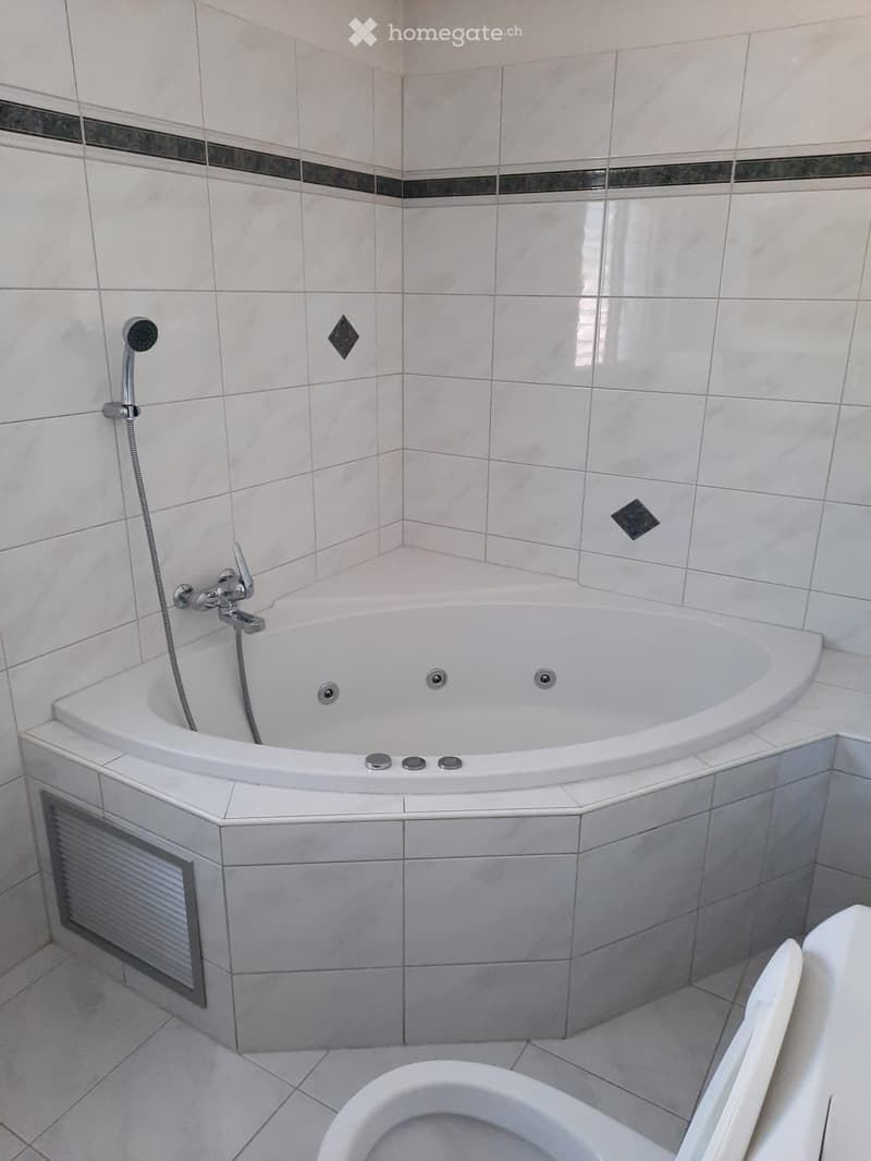 Modern bathroom with a white bathtub, chrome faucet, and a toilet. The walls are tiled with white tiles and decorative black diamond-shaped tiles.