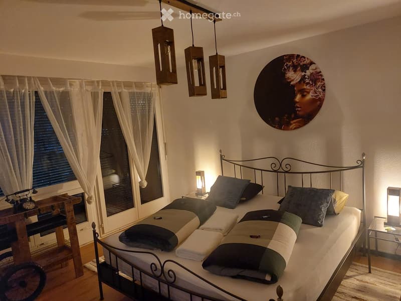 Cozy bedroom with a metal-framed bed, multiple pillows and blankets, nightstands with lamps, large window with curtains, and decorative wall art.