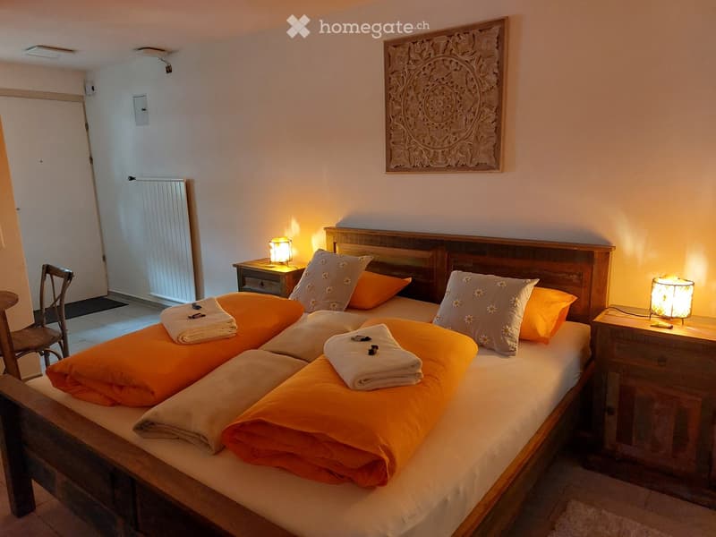 Cozy bedroom with a large wooden bed, orange blankets, decorative pillows, nightstands with lamps, a wooden chair, and a radiator near the door.