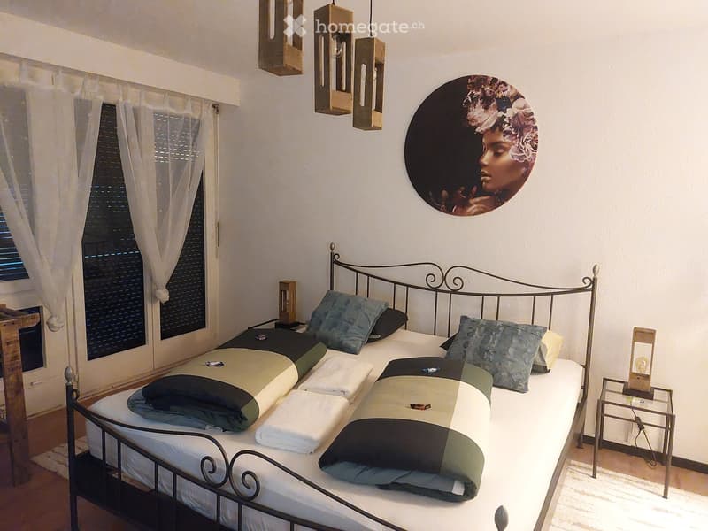 A well-lit bedroom with a metal-framed bed, multiple pillows, and a nightstand with a lamp. The room features large windows with sheer curtains and a decorative wall art piece.