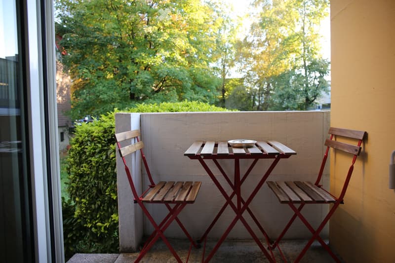 Balcony with a small red metal table and two matching chairs, set against a backdrop of greenery and a yellow wall.