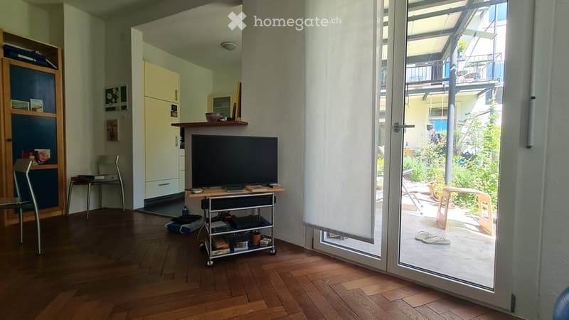 Modern living room with hardwood floors, large glass door leading to a balcony, TV stand with flat-screen TV, small table, chairs, and a cabinet with shelves.