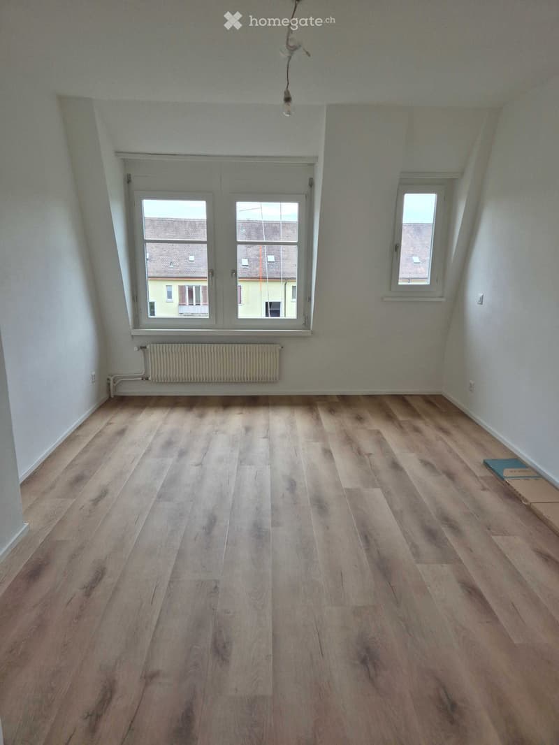 Empty room with wooden flooring, white walls, and large windows showing a view of neighboring buildings.