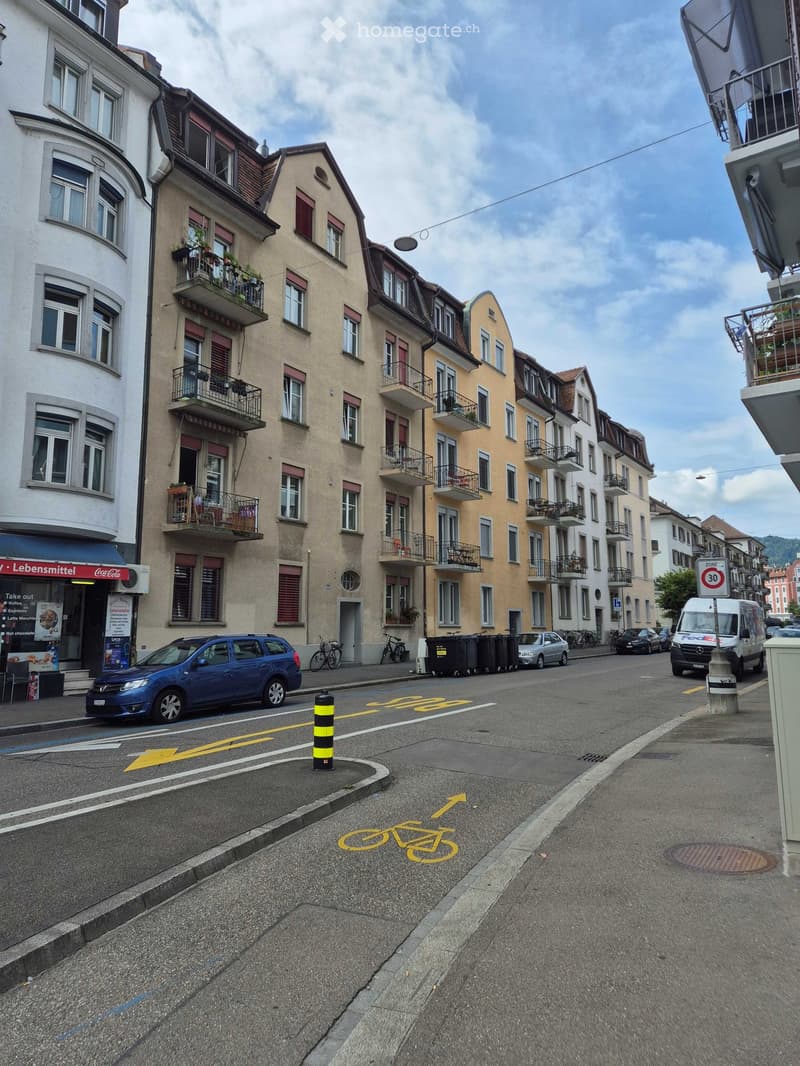 A street lined with multi-story residential buildings featuring balconies, windows, and a mix of architectural styles. The road includes a bicycle lane, parked cars, and a speed limit sign.