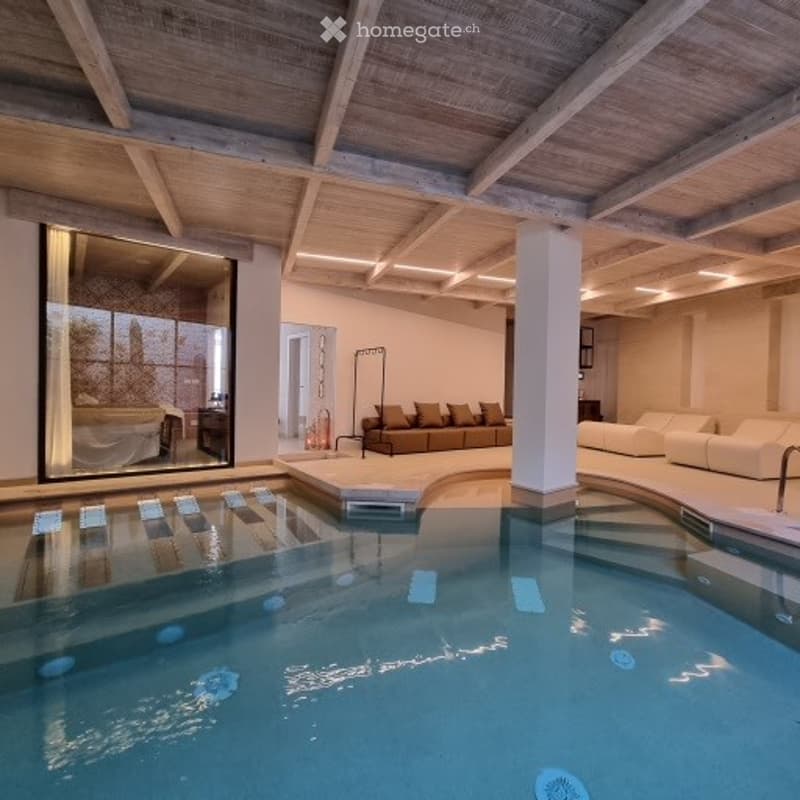 Indoor-outdoor pool area with wooden beams, lounge seating, and a view of a bedroom through a large glass wall.