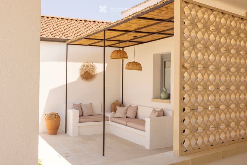 Outdoor patio with a white built-in sofa, decorative vases, and a decorative wall piece. The area is covered by a pergola with hanging wicker lamps.