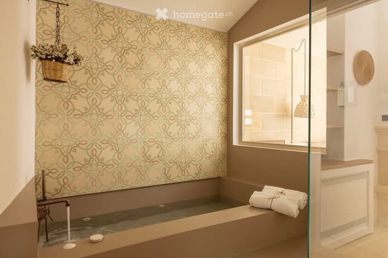 Bathroom with a large bathtub, decorative patterned wallpaper, a hanging wicker basket with flowers, a glass shower enclosure, and neatly folded towels on the edge of the tub.