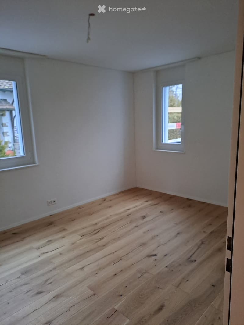 Empty room with wooden floor, two windows, white walls, a power outlet