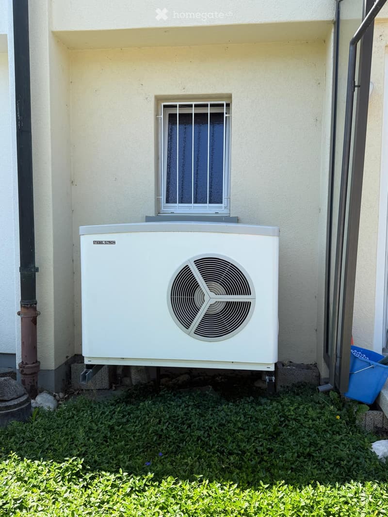 A white air conditioning unit is mounted on a wall with a small window above it. The surrounding area is covered with green grass and small plants.